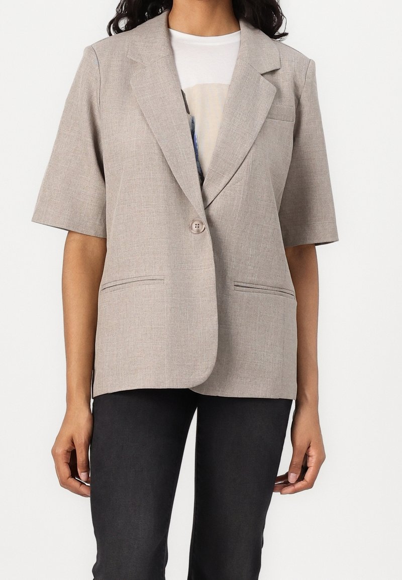 Femme portant un blazer beige à manches courtes sur un t-shirt blanc et un pantalon noir, montrée du cou jusqu'aux hanches sur un fond uni.