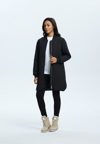 Veste longue noire matelassée avec fermeture éclair sur le devant, col rond et poches latérales, assortie à des leggings noirs et des bottes d'hiver beiges doublées de fourrure.