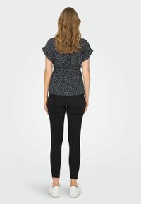 Blouse noire à imprimé floral avec manches à volants, taille froncée, associée à un jean skinny noir et des baskets blanches. Vue arrière visible.