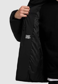 Doudoune noire avec un intérieur lisse et brillant. Dispose d'une poche zippée sur le côté et d'un patch logo à l'intérieur. Design classique avec un col haut.