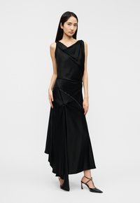 Abito in raso nero con scollo drappeggiato, orlo asimmetrico e cuciture decorative. Abbinato a scarpe nere con tacco e accessori minimalisti.