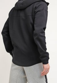 Person med svart huva jacka och ljusgråa sweatpants med svart rand, stående med ryggen vänd mot kameran.