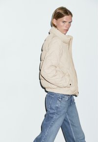 Beige gewatteerde pufferjack met een hoge kraag, ritsluiting en elastische manchetten, gecombineerd met lichtblauwe straight-leg jeans.