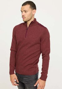 Pull à zip demi hauteur bordeaux avec texture côtelée et col haut. Présente des accents de couleur maroon contrastants sur les épaules. Porté avec un jean foncé.