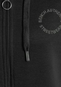 Haut à capuche zippé noir avec une texture lisse, cordon tressé avec embouts en métal et logo brodé "Bench Authentic Streetwear".