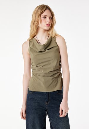 Ung kvinde med bølget blondt hår iført en olivengrøn ærmeløs top og mørkeblå jeans, der kigger mod højre mod en hvid baggrund.