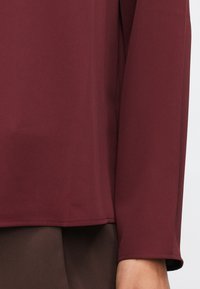 Μαρονάκι μακρυμάνικο πουκάμισο με μανίκι και στρίφωμα πάνω από καφέ παντελόνι, δείχνοντας μέρος του χεριού και του χεριού ενός ατόμου σε ουδέτερο φόντο.