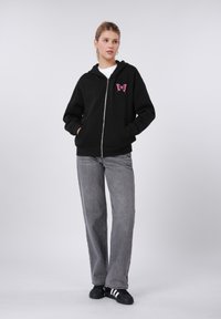 Sweat à capuche noir zippé avec un motif papillon rose sur la poitrine, accompagné d'un pantalon gris à jambes larges et de chaussures noires avec des rayures blanches.