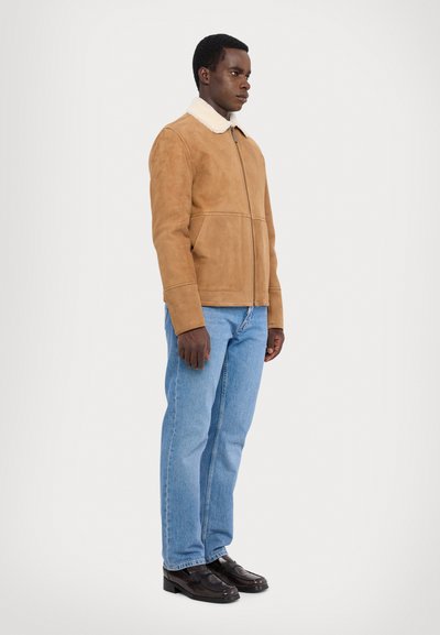 Tan ruskindsjakke med shearlingkrave, frontlynlås og sidelommer, parret med lyseblå straight-leg jeans og sorte sko.