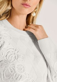 Frau trägt einen weißen strukturierten Strickpullover mit floralen Spitzen-Details und großen goldenen sowie Perlen-Ohrringen, Hand in der Nähe des Halsbereichs.