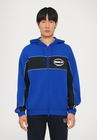 Plava zip-up dukserica s crno-bijelim prugama, s logom na prsima i rebrastim manžetama. Mekani materijal s opuštenim krojem.