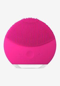 Dispositif de nettoyage facial en silicone rose, en forme ovale avec des brins de silicone texturés sur le dessus, boutons de contrôle plus/moins, et une base blanche.