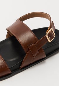 Sandalia de cuero marrón con dos correas anchas, una plantilla texturizada negra y una hebilla ajustable en la correa del tobillo.