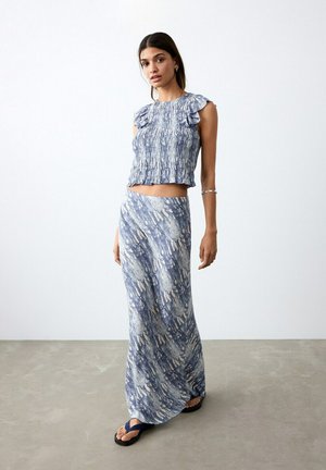 Žena stoji, nosi plavi i bijeli uzorak na majici bez rukava s volanima i usklađenu dugu suknju, uz crne sandale.