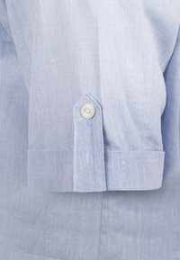 Camisa de lino azul claro con un tejido texturizado, cierre de un botón en los puños de las mangas y un diseño de puño puntiagudo.