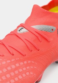 Jasny koralowy but piłkarski z teksturowaną powierzchnią, cienkimi sznurowadłami i odblaskowymi akcentami. Charakteryzuje się dopasowanym kształtem i niskoprofilowym designem.