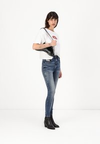 Mršave plave traperice, visokog struka, blago istrošene, uparene s bijelom majicom s crvenim logom. Crna torba preko ramena i gležnjače.