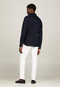 Camicia a maniche lunghe blu navy con colletto a punta e polsini con bottone, abbinata a pantaloni bianchi e scarpe slip-on blu navy. Tessuto liscio, vestibilità sagomata.