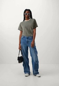 Woman with braided hair wearing white sunglasses, black-and-white striped t-shirt, blue cargo jeans, white sneakers, holding a black handbag.