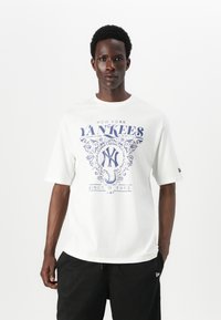 Baltas medvilnės marškinėliai su mėlyna "New York Yankees" grafika, kuri turi dailius ornamentus ir metus "1903". Atsipalaidavęs siluetas su trumpomis rankovėmis.