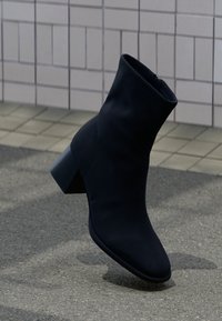 Μαύρο μεσαίο μποτάκι με τακούνι που φτάνει μέχρι τη μέση της γάμπας, στέκεται πάνω σε γκρι πεζοδρόμιο μπροστά από πλακιδωτό τοίχο, πλαϊνή όψη με τετράγωνο τακούνι και στρογγυλεμένη μύτη.