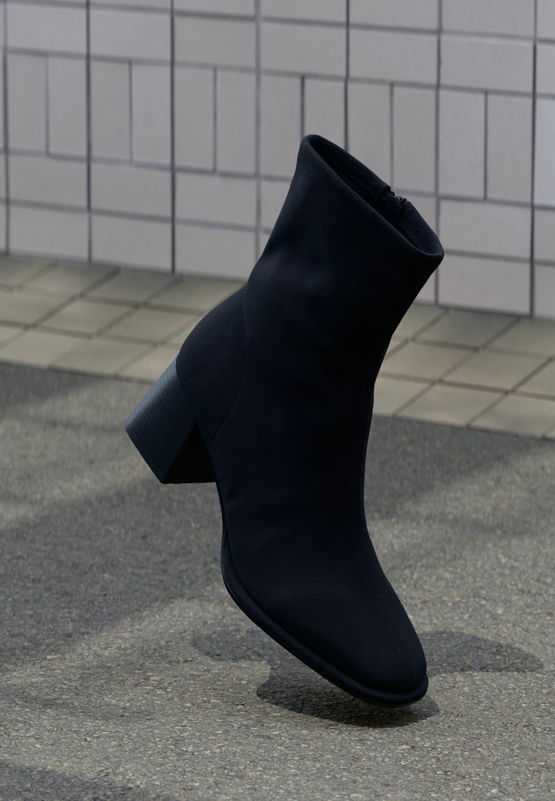 Μαύρο μεσαίο μποτάκι με τακούνι που φτάνει μέχρι τη μέση της γάμπας, στέκεται πάνω σε γκρι πεζοδρόμιο μπροστά από πλακιδωτό τοίχο, πλαϊνή όψη με τετράγωνο τακούνι και στρογγυλεμένη μύτη.