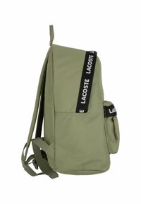 Mochila de tela verde con una forma estructurada, correa con logotipo negro, bolsillo lateral y tirantes ajustables, diseñada para la funcionalidad y el estilo.