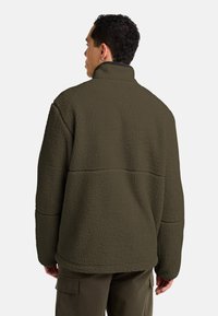 Dark green fleece jacket with a high collar, textured surface, and relaxed fit. Features contrasting stitching and dropped shoulder seams.