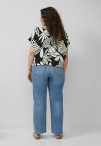 Schwarzes Oberteil mit weißen Blattmustern, kurze Ärmel, kombiniert mit hellblauen geraden Jeans und beigen Sandalen. Rückansicht.