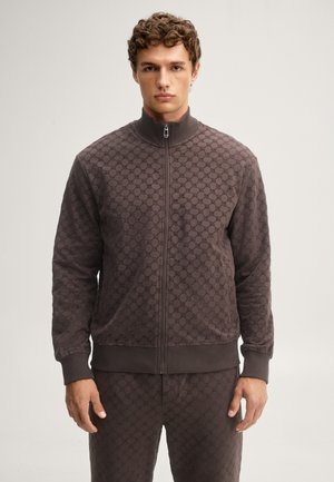 Jonge man met krullend haar die een donkerbruine jas met patroon en rits draagt en een bijpassende broek, staand tegen een effen lichte achtergrond.