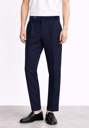 Mężczyzna noszący granatowe, dopasowane spodnie z mankietami i czarne skórzane loafersy, stojący na szarej podłodze na tle białej ściany.