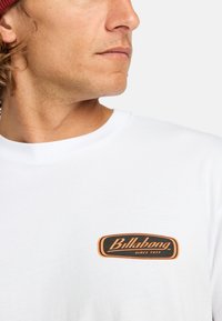 Hvid bomulds t-shirt med et sort og orange "Billabong" logo-patch på venstre bryst. Korte ærmer og rund halsudskæring.
