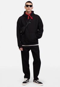 Un sweat à capuche noir avec une poche avant, associé à un pantalon noir. Accessoirisé avec un bandana rouge et un sac bandoulière noir. Des baskets noires avec des semelles blanches.