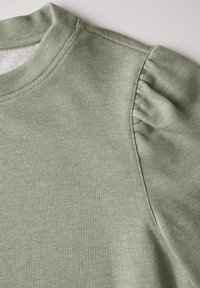 Grön bomullströja med en mjuk textur, rund halsringning och rynkad detalj på axeln. Sömmen är prydlig och jämn.