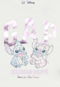 Δύο καρτούν χαρακτήρες Stitch τρώνε χωνάκια παγωτού κάτω από λαμπερό ροζ κείμενο "GAP" και παστέλ κείμενο "ICE CREAM SHOPPE here for the treats" σε λευκό ύφασμα.