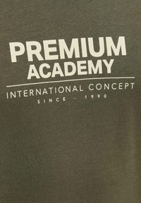 Olivno zelena bombažna majica z bold belim napisom: "PREMIUM ACADEMY," "INTERNATIONAL CONCEPT" in "SINCE - 1990" v preprostem postavitvi.