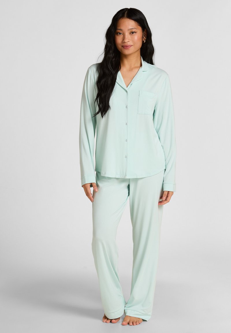 Frau mit langen dunklen Haaren, die ein hellgrünes Pyjama-Set mit Knöpfen trägt und barfuß vor einem einfarbigen Hintergrund steht.