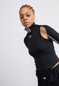 Young person with braided hair wears black Adidas athletic wear with cutout sleeves and logo, posing against plain light background.