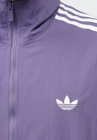 Lilla lukuga jakk kõrge kraega, millel on õlgadel kolm valget triipu ja rinnal valge kolmleheline logo.