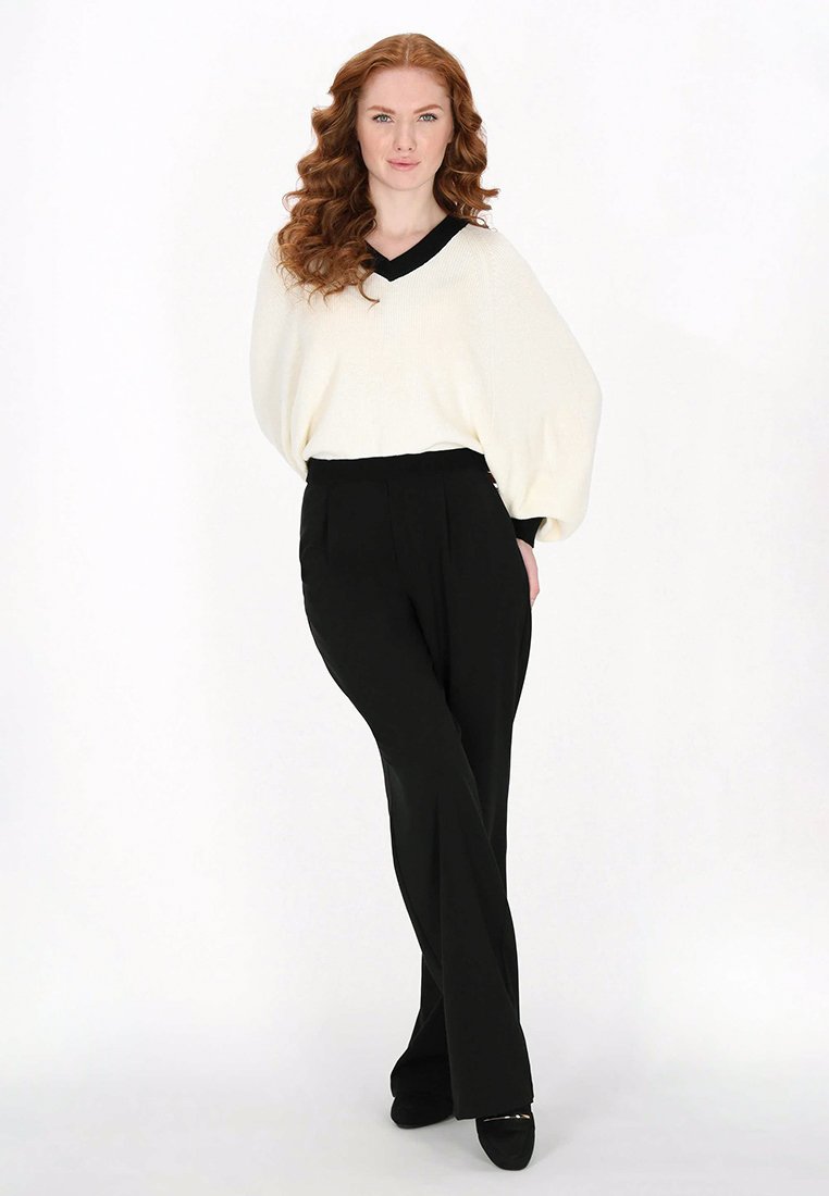 Weißer oversized Pullover mit einem schwarzen V-Ausschnitt, kombiniert mit hochtaillierten schwarzen Hose. Der Pullover hat ein strukturiertes Strickmuster und eine lässige Passform.