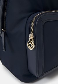 Zaino in nylon blu navy con dettagli in cerniera dorati e un tirante decorativo. Presenta un design strutturato e una consistenza morbida.