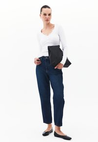 Blusa blanca de manga larga con detalles de encaje en el escote, jeans de cintura alta azules y zapatos planos negros, sosteniendo un clutch de cuero negro.