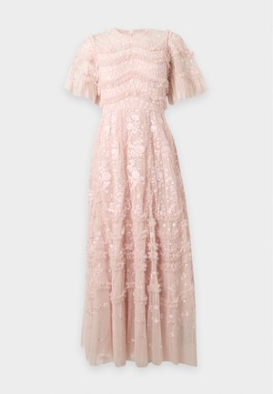 Femme portant une robe maxi en dentelle florale rose pâle avec de courtes manches à volants et des talons ouverts beige, debout devant un fond blanc.