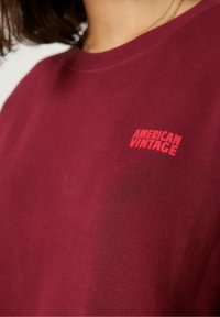 Bordo spalvos džemperis, turintis apvalų iškirpimą ir trumpas rankoves, pagamintas iš minkštos medžiagos su mažu raudonu "American Vintage" logotipo detaliu.