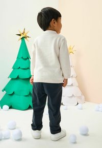 Ein Kind in einem hellbeigen Pullover und dunkelblauen Hosen steht neben grünen und weißen Papier-Weihnachtsbäumen, umgeben von weißen Pompons auf einem weißen Boden.