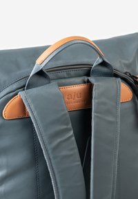 Grauer Stoffrucksack mit Lederakzenten, ausgestattet mit gepolsterten Trägern und einem oberen Griff. Enthält ein gebrandetes Lederetikett zum Beschriften.