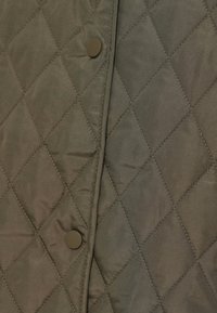 Πράσινο υφασμάτινο κουβέρτα με διαμάντι μοτίβο· δύο καφέ κουμπιά με κλίπ στο μπροστινό μέρος. Λεία υφή με διακριτική λάμψη.