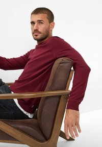 Bordeauxfarbener Pullover mit rundem Ausschnitt, glatter Textur, über einem weißen Hemd getragen, kombiniert mit dunkler Denim, sitzt in einem Holzsessel.