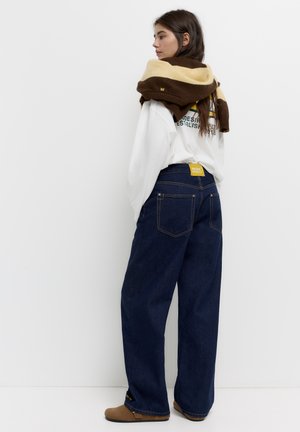 Široke tamnoplave traperice s leđnim džepovima, u kombinaciji s bijelom majicom dugih rukava i smešanožutom prugastom pletenom maramom.