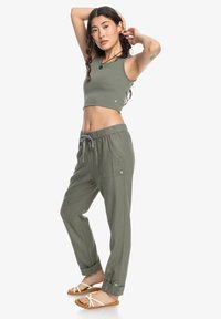 Žena v olivovo zelenej crop top a voľných nohaviciach sťahovacím pásom pózuje bosá na bielej pozadí s rukami zdvihnutými za hlavu.